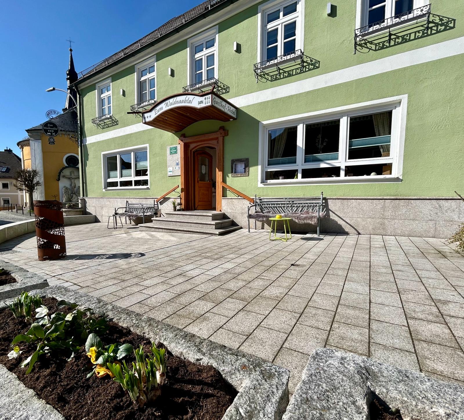 Eingangsbereich zum Hotel "Zum Waldnaabtal", davor ein gepflasterter Platz mit Sitzbänken, Frühlingsblumen